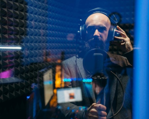 Audio Branding Soundtrack A bearded man in a recording studio is thoroughly absorbed in audio branding, wearing headphones and speaking into a microphone, surrounded by soundproof panels.