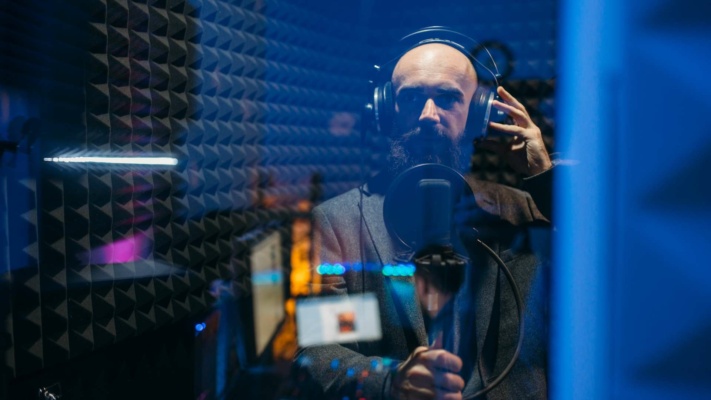 A bearded man in a recording studio is thoroughly absorbed in audio branding, wearing headphones and speaking into a microphone, surrounded by soundproof panels.