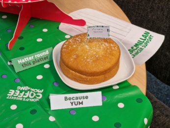 A Victoria sponge cake rests on a white plate, decorated with labels reading Victoria Sponge and Because YUM. A touch of coffee enhances the delightful scene, set on a green polka dot tablecloth prominently featuring Macmillan Cancer Support branding.