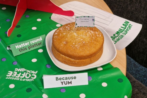 A Victoria sponge cake rests on a white plate, decorated with labels reading Victoria Sponge and Because YUM. A touch of coffee enhances the delightful scene, set on a green polka dot tablecloth prominently featuring Macmillan Cancer Support branding.