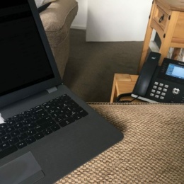 Laptop and office telephone on a textured brown surface in a cosy work-from-home setting, complete with a sofa and wooden side table.