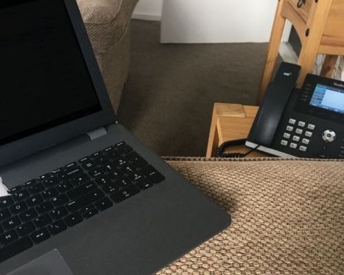 Our phone solutions can transform the way you work and where you work Laptop and office telephone on a textured brown surface in a cosy work-from-home setting, complete with a sofa and wooden side table.
