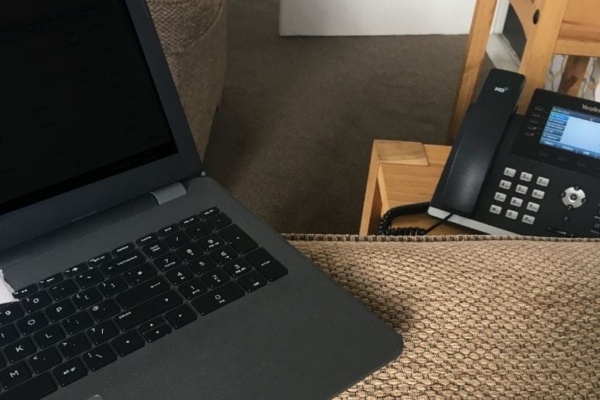 Laptop and office telephone on a textured brown surface in a cosy work-from-home setting, complete with a sofa and wooden side table.