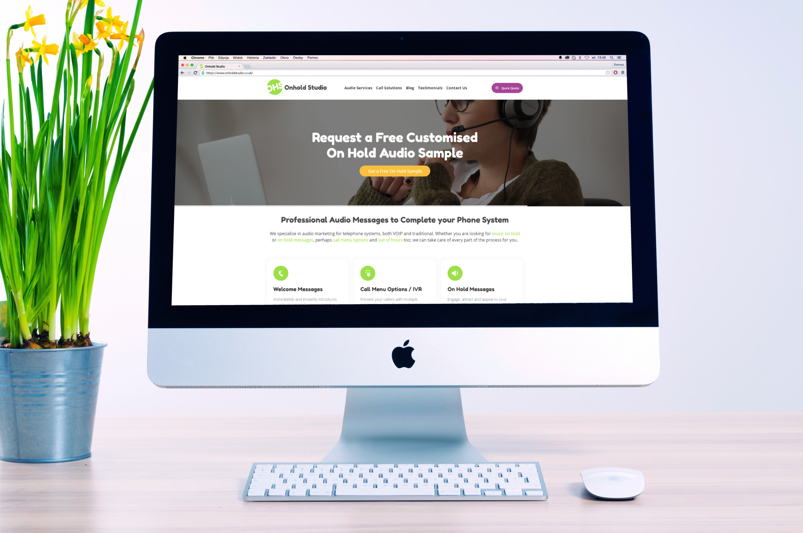 A computer monitor displays a website offering free customised on-hold audio samples, perfect for enhancing your on-hold marketing strategy. A keyboard and mouse sit below the screen, with a potted plant adding a touch of greenery on the left.