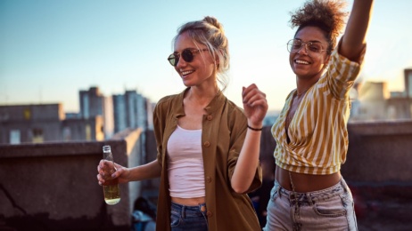 Two women stand on a rooftop at sunset, smiling and dancing; one holds a bottle while both wear sunglasses and casual clothes. City buildings are visible in the background.