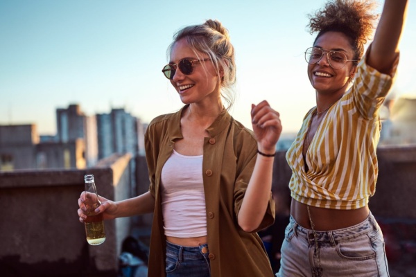 Two women stand on a rooftop at sunset, smiling and dancing; one holds a bottle while both wear sunglasses and casual clothes. City buildings are visible in the background.
