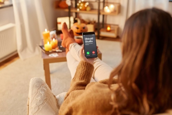 A person in cosy clothing sits on a sofa holding a mobile with an incoming call from “Onhold Studio”; candles and autumn decorations are visible in the background.