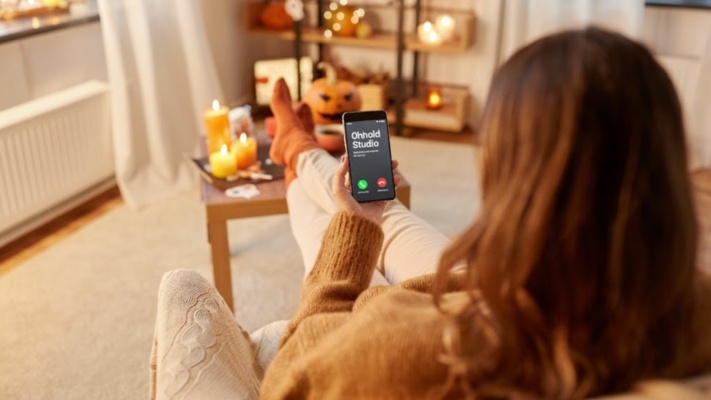 Autumn Royalty Free Music A person in cosy clothing sits on a sofa holding a mobile with an incoming call from “Onhold Studio”; candles and autumn decorations are visible in the background.
