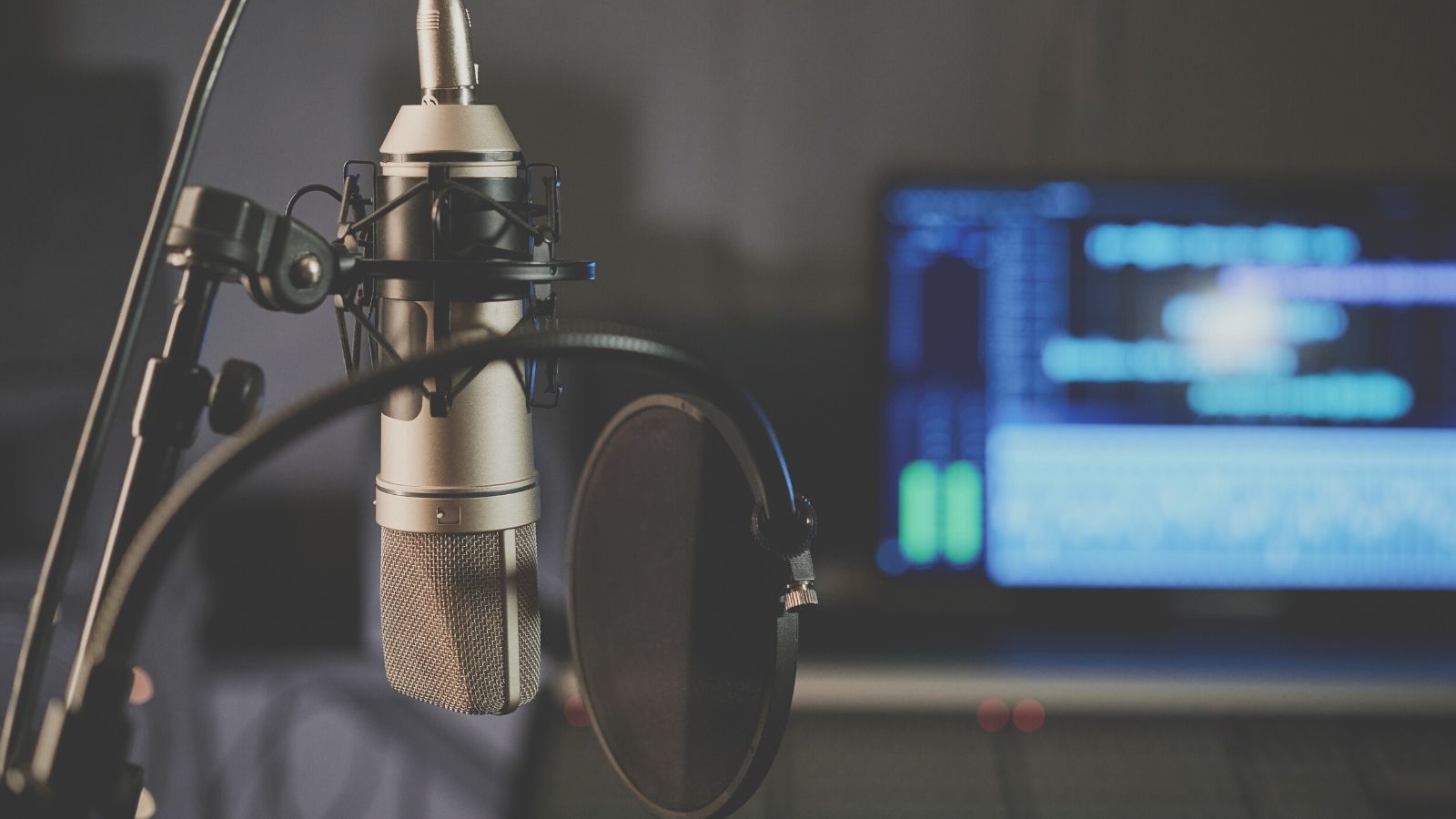 A studio microphone with a pop filter in front, set against a blurred background showing a computer screen with audio editing software used by an on-hold provider.