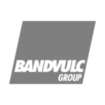 bandvulc-group A person in a checked shirt kneels at a riverbank surrounded by tall plants. They look across the water, set against a forested landscape. The sky is overcast.