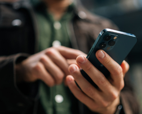 A person in a brown jacket and green shirt uses a mobile phone outdoors, concentrating on the screen—possibly conducting business phone communication with one hand whilst touching the screen with the other.