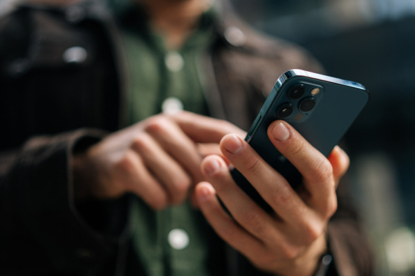 A person in a brown jacket and green shirt uses a mobile phone outdoors, concentrating on the screen—possibly conducting business phone communication with one hand whilst touching the screen with the other.