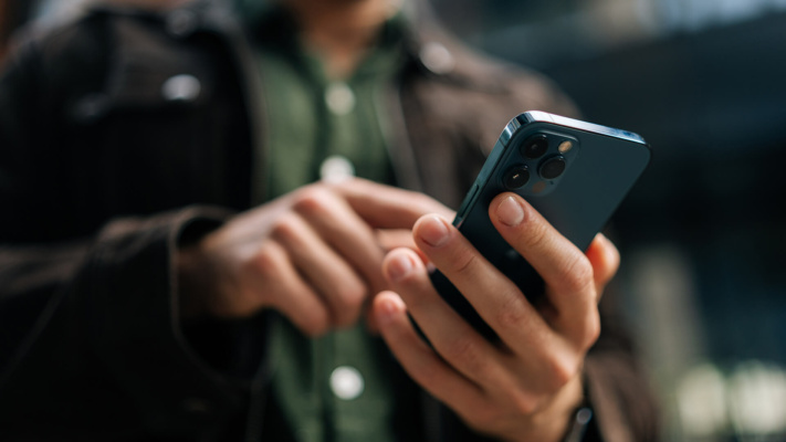 A person in a brown jacket and green shirt uses a mobile phone outdoors, concentrating on the screen—possibly conducting business phone communication with one hand whilst touching the screen with the other.