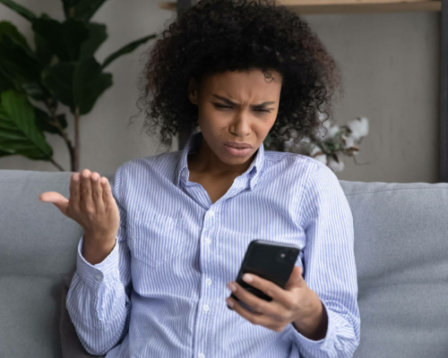 A person sitting on a settee looks at their mobile with a confused and frustrated expression, holding one hand up in disbelief.