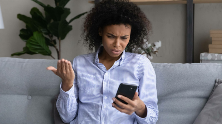 blog-annoyed-on-phone A person sitting on a settee looks at their mobile with a confused and frustrated expression, holding one hand up in disbelief.