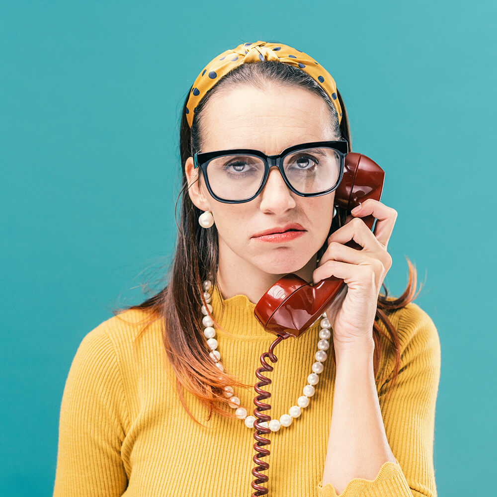 Woman in large spectacles and a yellow jumper holds a red rotary telephone to her ear, appearing frustrated against a teal background.