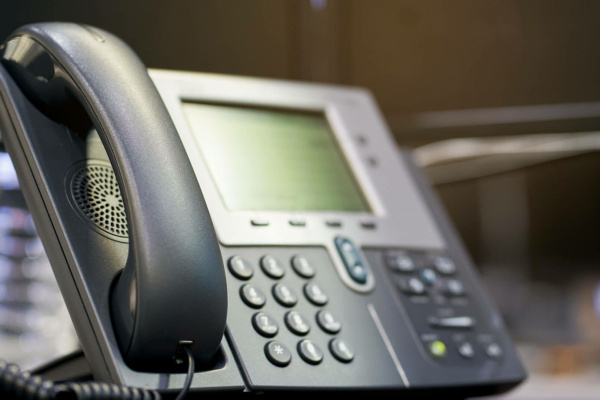 Close-up of an office desk telephone with a digital display screen, buttons, and a corded handset. The background is blurred.