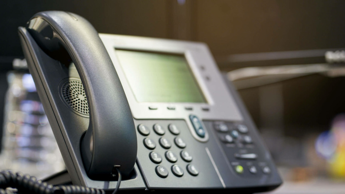 Close-up of an office desk telephone with a digital display screen, buttons, and a corded handset. The background is blurred.