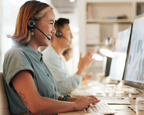 A smiling customer service agent wearing a headset types at a computer, with colleagues working in the background.