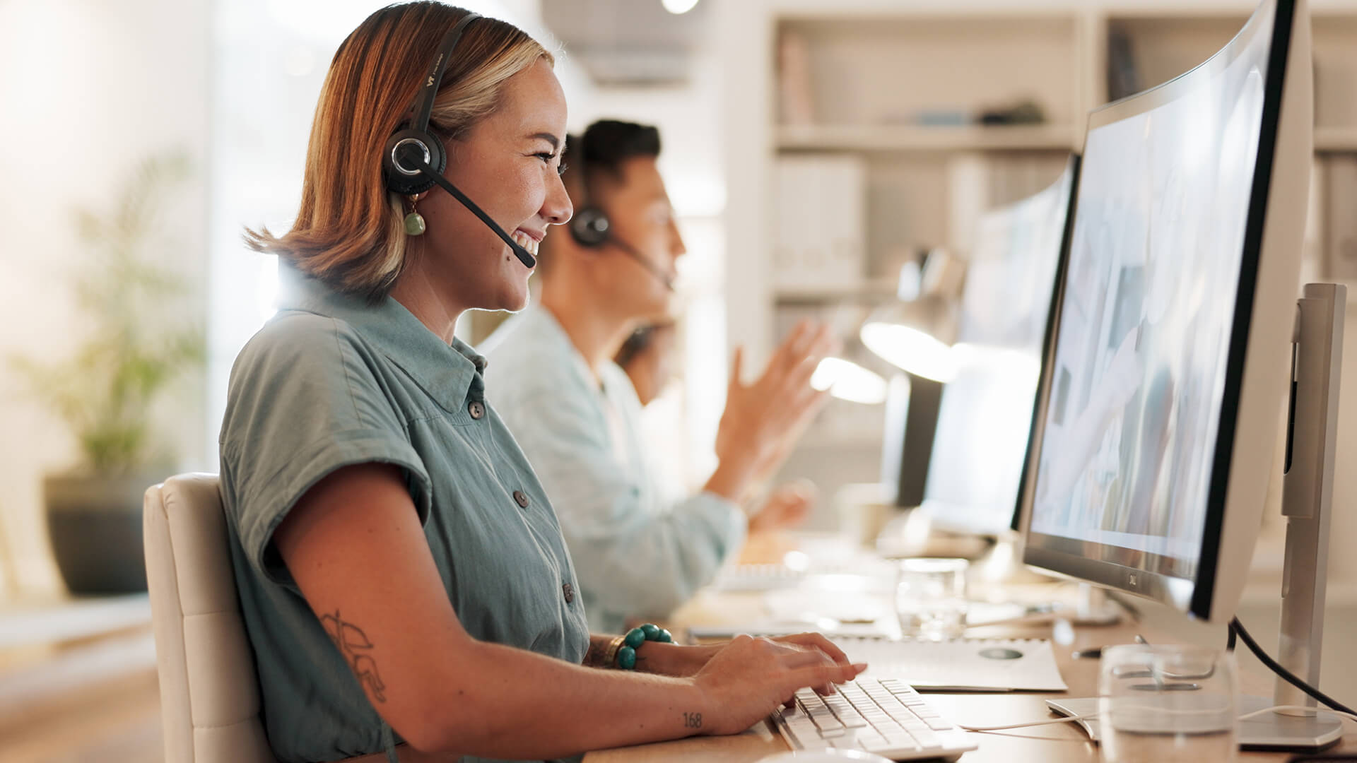 A smiling customer service agent wearing a headset types at a computer, with colleagues working in the background.