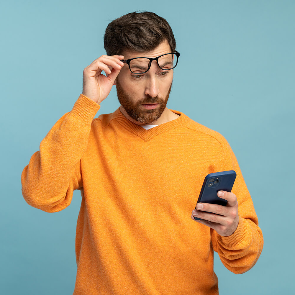A man in an orange jumper holds a mobile phone in one hand and adjusts his glasses with the other, looking at the phone with a surprised expression against a blue background.