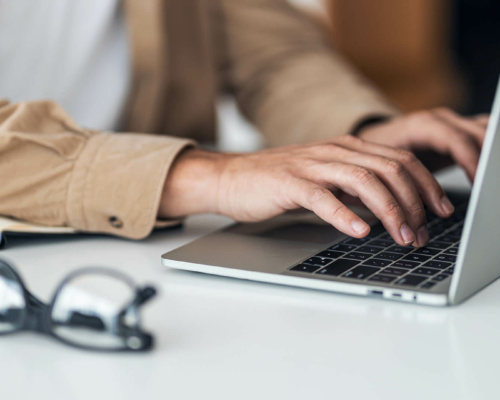 A person types on a laptop keyboard at a desk, with an open notebook and glasses nearby.