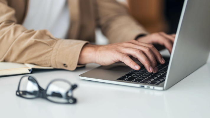 A person types on a laptop keyboard at a desk, with an open notebook and glasses nearby.