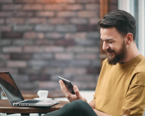 A bearded man sits at a café table with a laptop and coffee, smiling as he looks at his mobile.