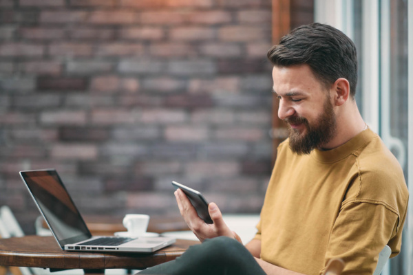 A bearded man sits at a café table with a laptop and coffee, smiling as he looks at his mobile.