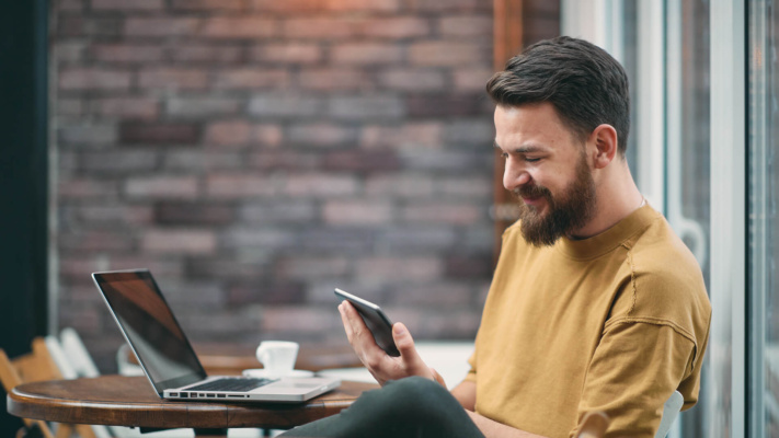 A bearded man sits at a café table with a laptop and coffee, smiling as he looks at his mobile.