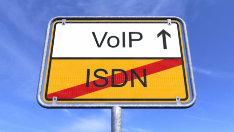 A road sign set against a blue sky displays “VoIP” with an upward arrow above and “ISDN” crossed out below, symbolising the PSTN switch-off and the shift from ISDN to VoIP technology.