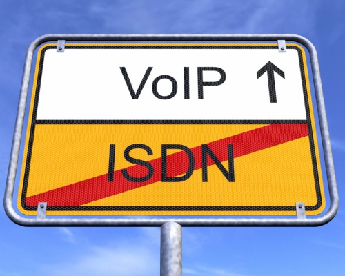 A road sign set against a blue sky displays “VoIP” with an upward arrow above and “ISDN” crossed out below, symbolising the PSTN switch-off and the shift from ISDN to VoIP technology.