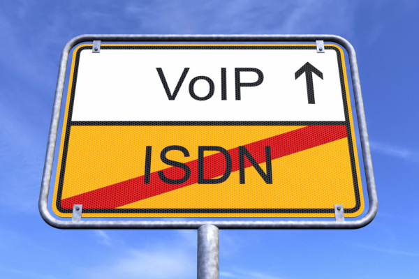 A road sign set against a blue sky displays “VoIP” with an upward arrow above and “ISDN” crossed out below, symbolising the PSTN switch-off and the shift from ISDN to VoIP technology.