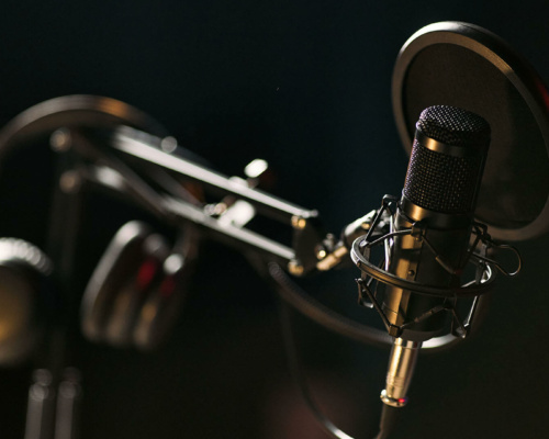 A black condenser microphone with a pop filter is mounted on a boom arm, with headphones hanging nearby, set against a dark, blurred background.