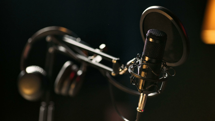 blog-microphone-closeup A black condenser microphone with a pop filter is mounted on a boom arm, with headphones hanging nearby, set against a dark, blurred background.