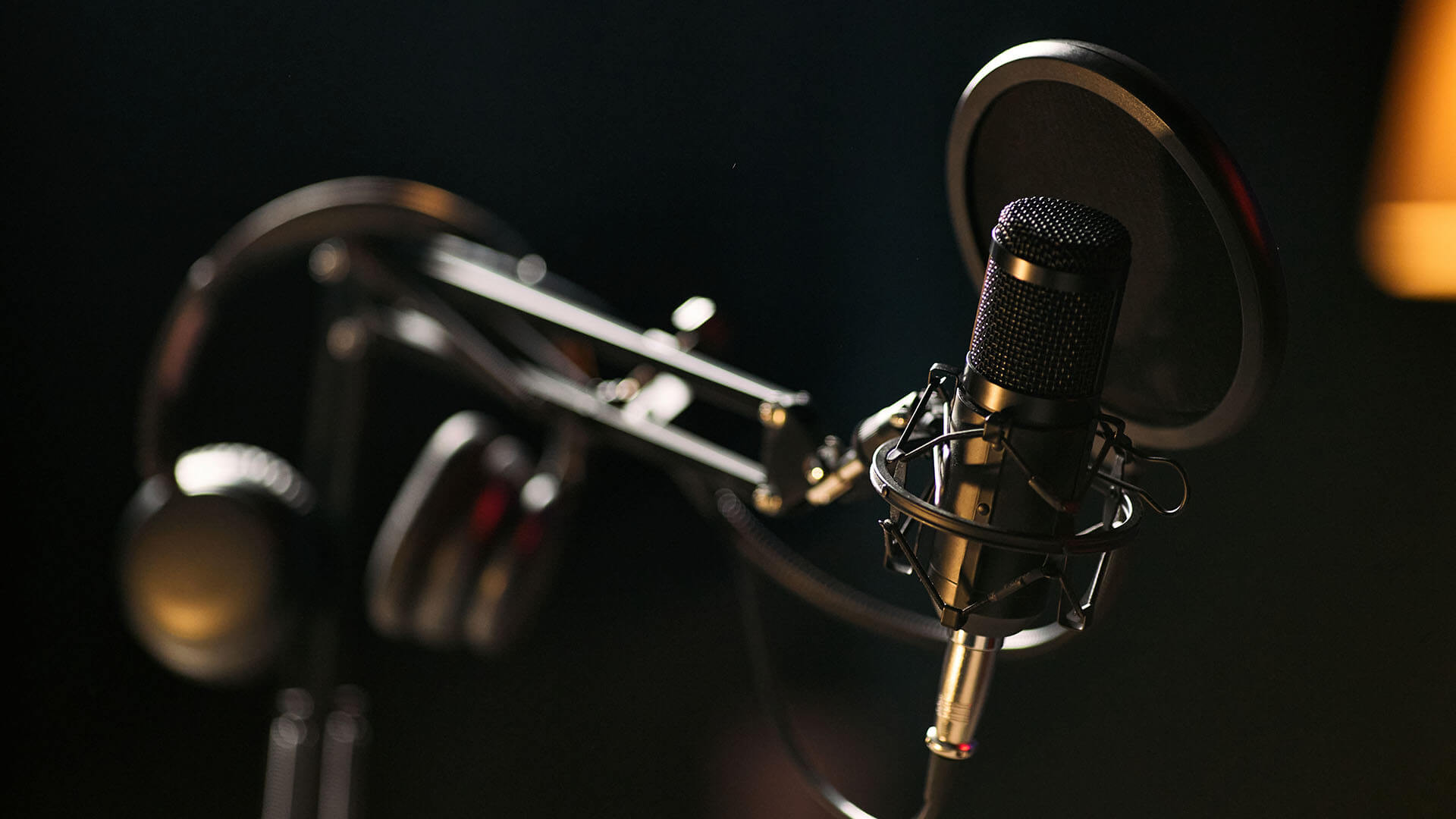 A black condenser microphone with a pop filter is mounted on a boom arm, with headphones hanging nearby, set against a dark, blurred background.