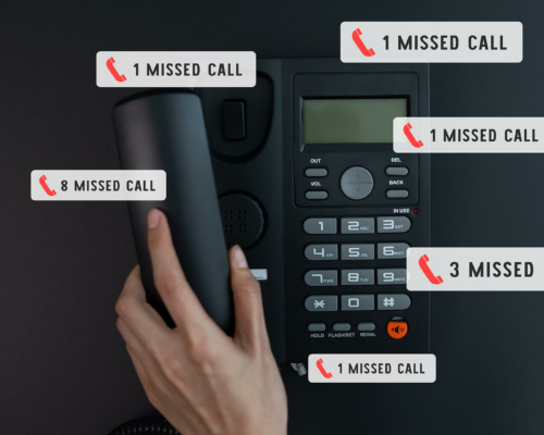 A person’s hand lifts the receiver of a black office phone displaying several missed call notifications with various numbers on a dark surface.
