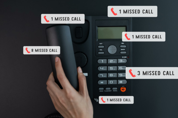 A person’s hand lifts the receiver of a black office phone displaying several missed call notifications with various numbers on a dark surface.