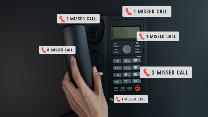 A person’s hand lifts the receiver of a black office phone displaying several missed call notifications with various numbers on a dark surface.