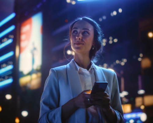 A woman in a light blazer holds a mobile phone and gazes upward, standing outdoors at night with bright city lights and illuminated hoardings in the background.