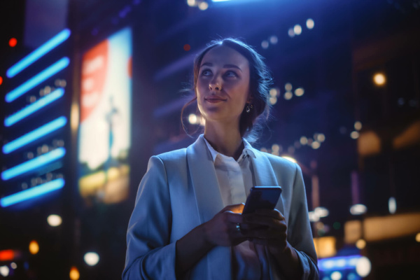 A woman in a light blazer holds a mobile phone and gazes upward, standing outdoors at night with bright city lights and illuminated hoardings in the background.