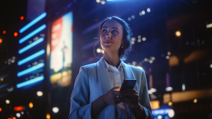 A woman in a light blazer holds a mobile phone and gazes upward, standing outdoors at night with bright city lights and illuminated hoardings in the background.