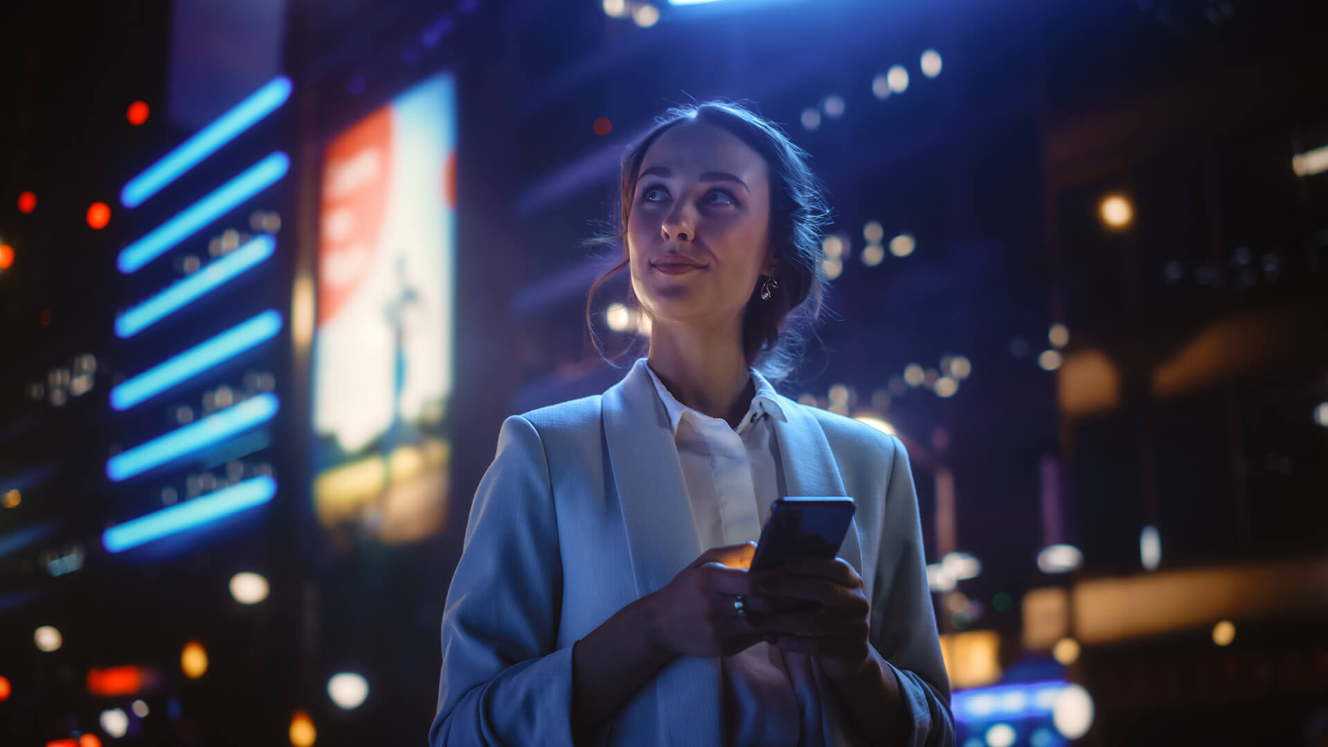 blog-mobile A woman in a light blazer holds a mobile phone and gazes upward, standing outdoors at night with bright city lights and illuminated hoardings in the background.