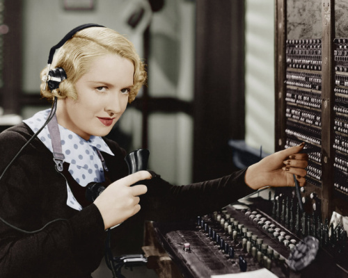 A blonde-haired woman operates a vintage telephone switchboard in an office, holding a handheld receiver and wearing a headset.
