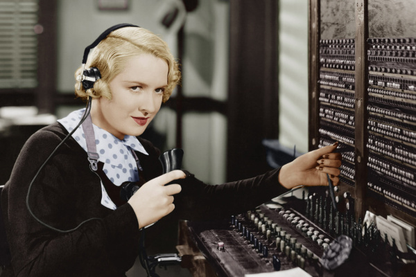 A blonde-haired woman operates a vintage telephone switchboard in an office, holding a handheld receiver and wearing a headset.