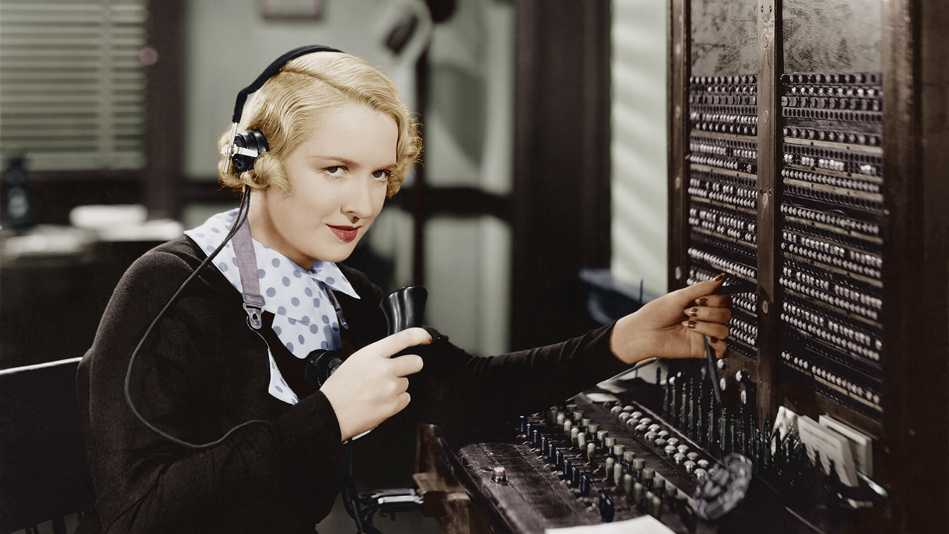 A blonde-haired woman operates a vintage telephone switchboard in an office, holding a handheld receiver and wearing a headset.