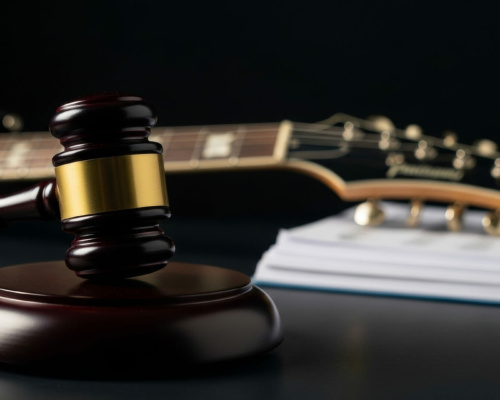 A judge’s gavel rests before a guitar and a pile of documents on a dark surface, representing legal issues concerning music or intellectual property.