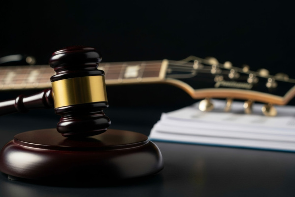 A judge’s gavel rests before a guitar and a pile of documents on a dark surface, representing legal issues concerning music or intellectual property.