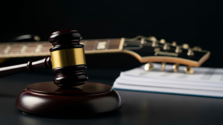 A judge’s gavel rests before a guitar and a pile of documents on a dark surface, representing legal issues concerning music or intellectual property.
