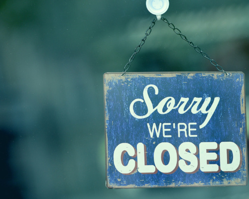 A blue and white sign hanging on a glass door reads Sorry We’re Closed.